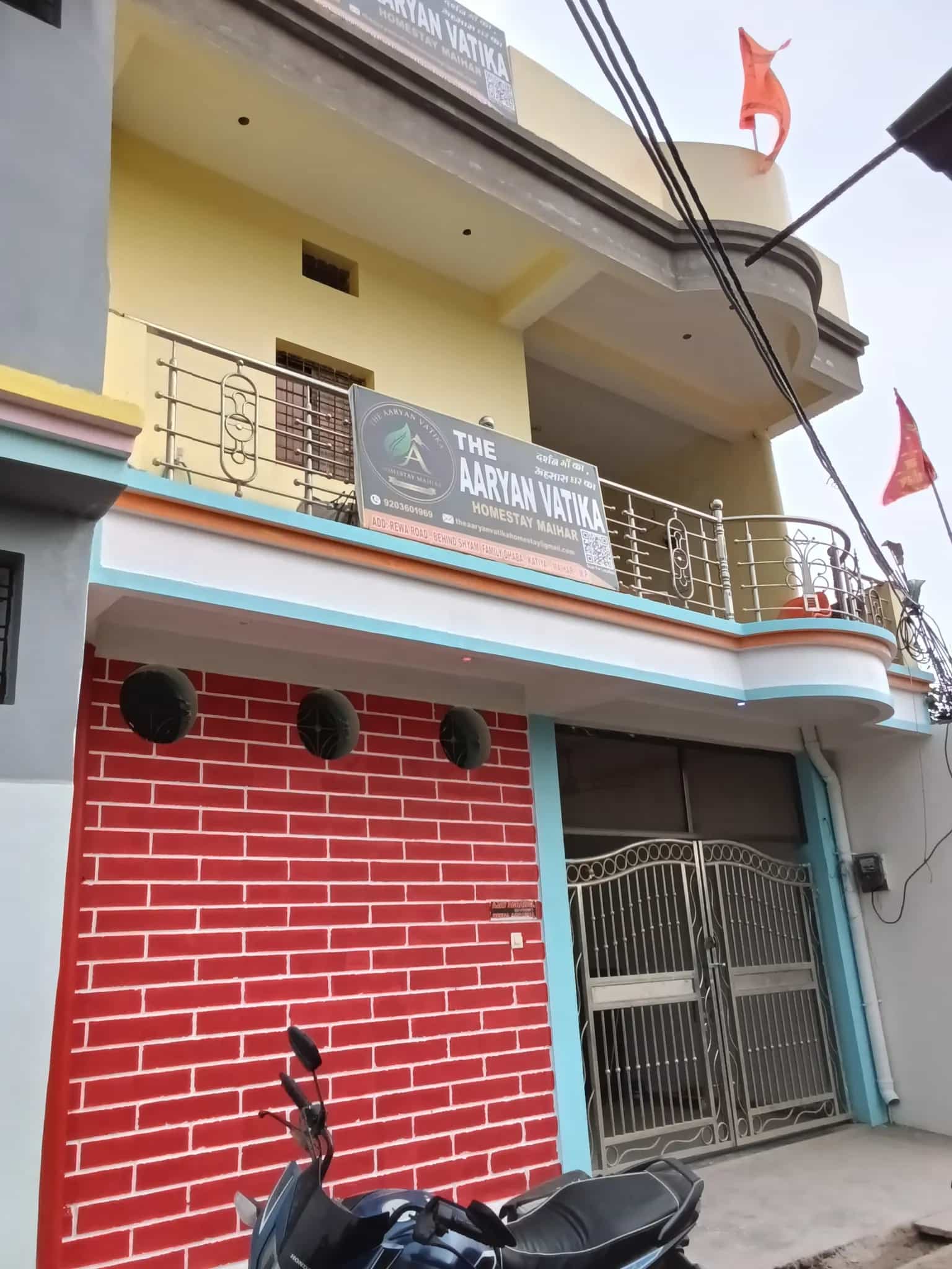 Exterior of a multi-story homestay in light yellow and gray, with a balcony, red brick wall, and a motorcycle.